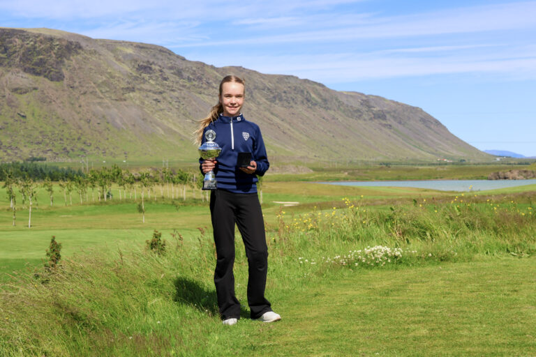 Young female golfer stands on a green field holding a large trophy, mountain backdrop and blue sky behind her.