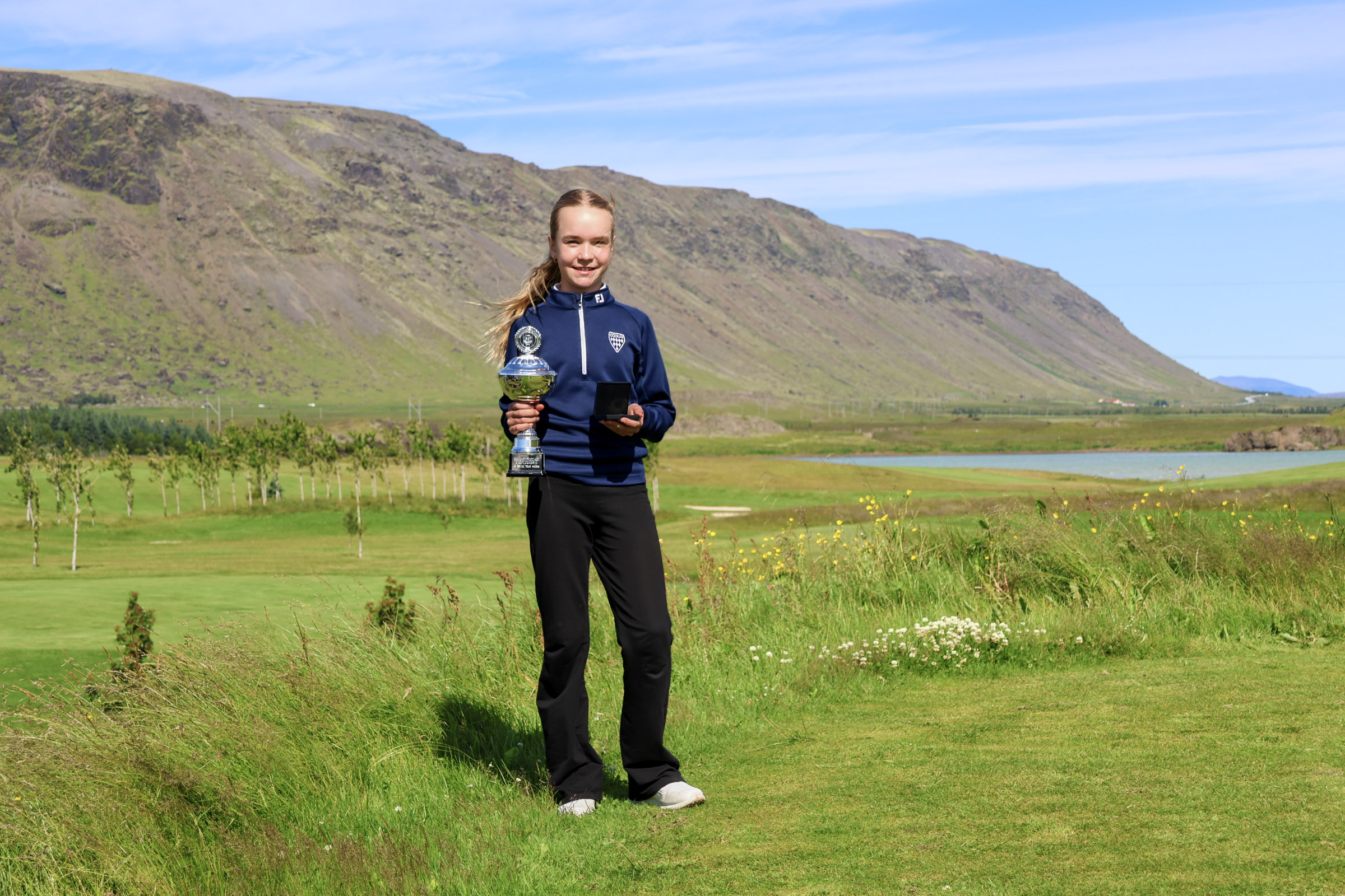 Young female golfer stands on a green field holding a large trophy, mountain backdrop and blue sky behind her.