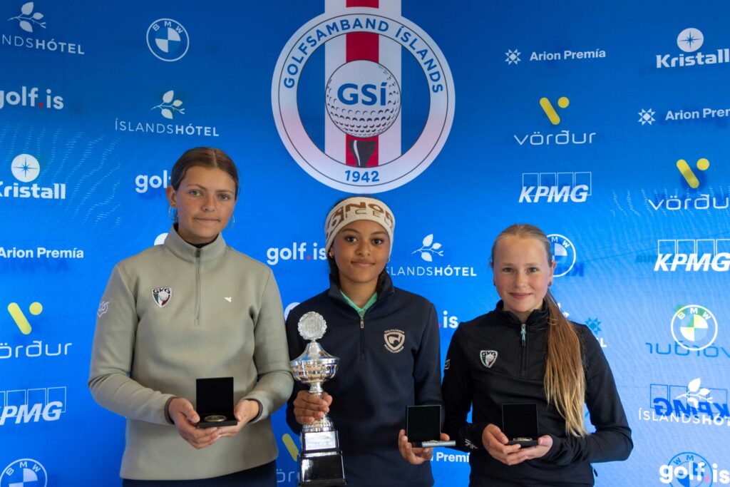 499A4030 2 - Golfsamband Íslands Three young female athletes stand on a podium holding trophies against a blue sponsor backdrop.