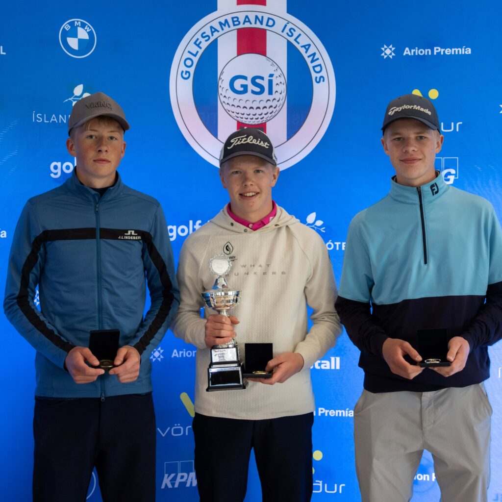 499A4057 1 - Golfsamband Íslands Three young men stand on a blue backdrop podium; the center figure holds a trophy while the other two hold medals, suggesting a sports award ceremony.