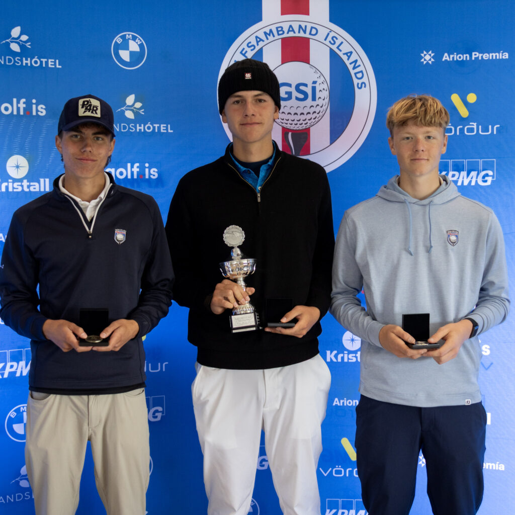 499A4073 1 - Golfsamband Íslands Three athletes stand side by side, each holding a trophy, against a blue sponsor backdrop.”,