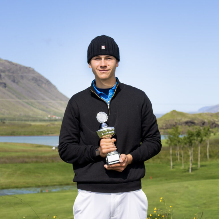 Young man in a black knit cap and jacket holding a silver trophy outdoors, with hills and a lake in the background.