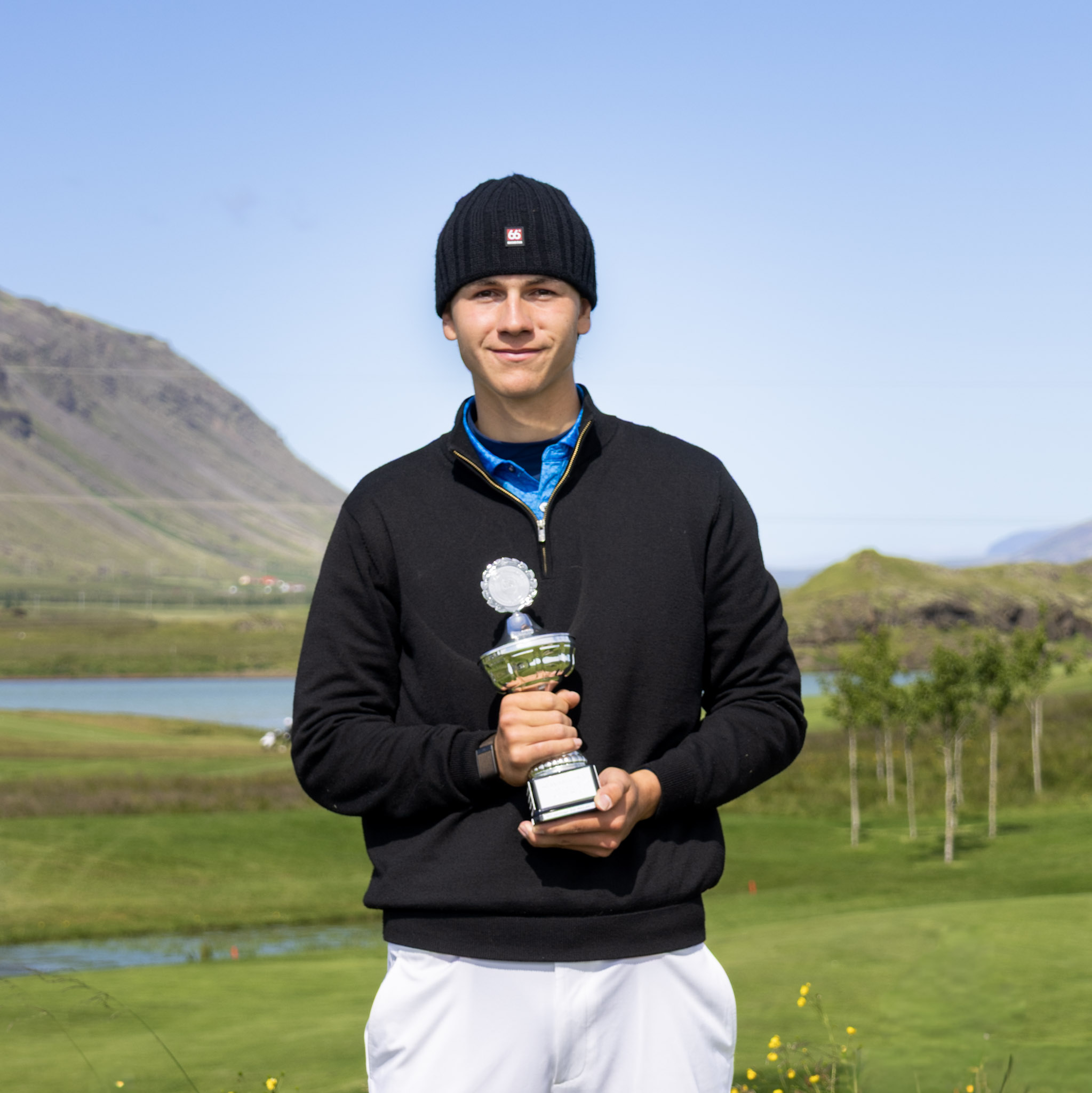 Young man in a black knit cap and jacket holding a silver trophy outdoors, with hills and a lake in the background.