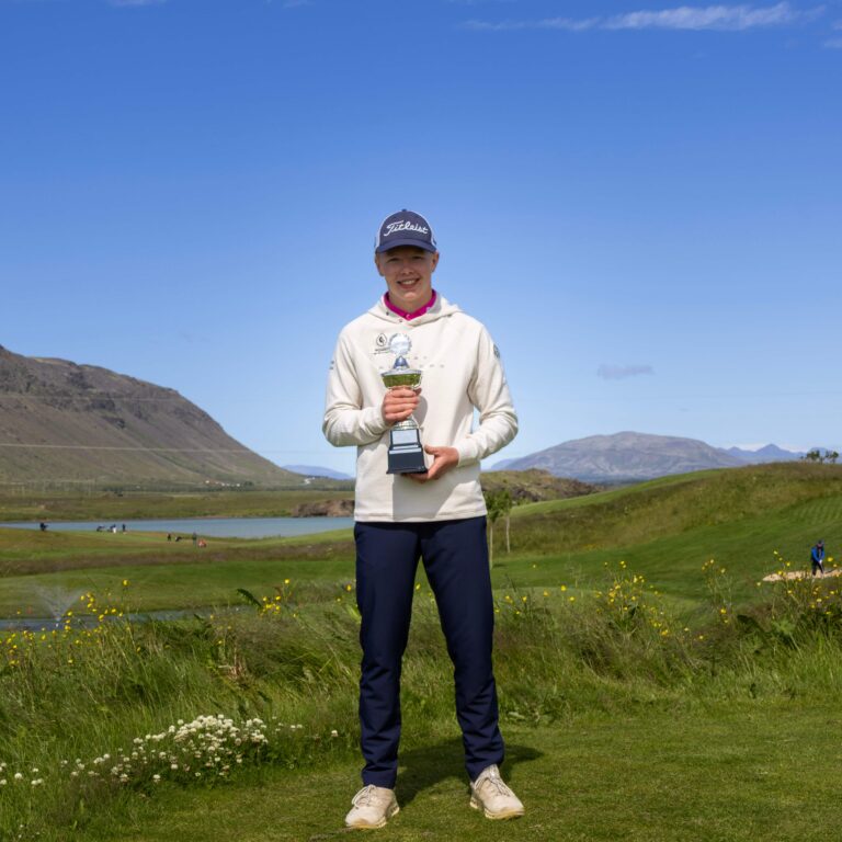 Young man standing on a grassy hilltop, holding a trophy, wearing a Titleist cap and white hoodie, with mountains and a clear blue sky behind him.
