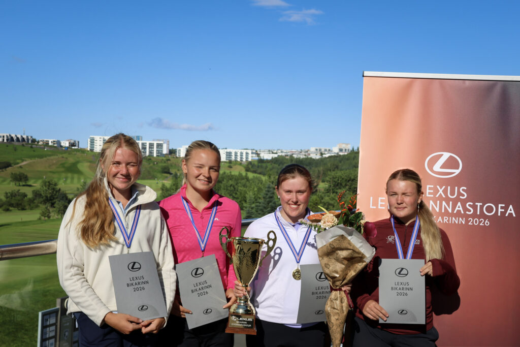 Four female athletes pose on a winners' podium with medals, a trophy, and bouquets outdoors against a blue sky and sponsor banner.