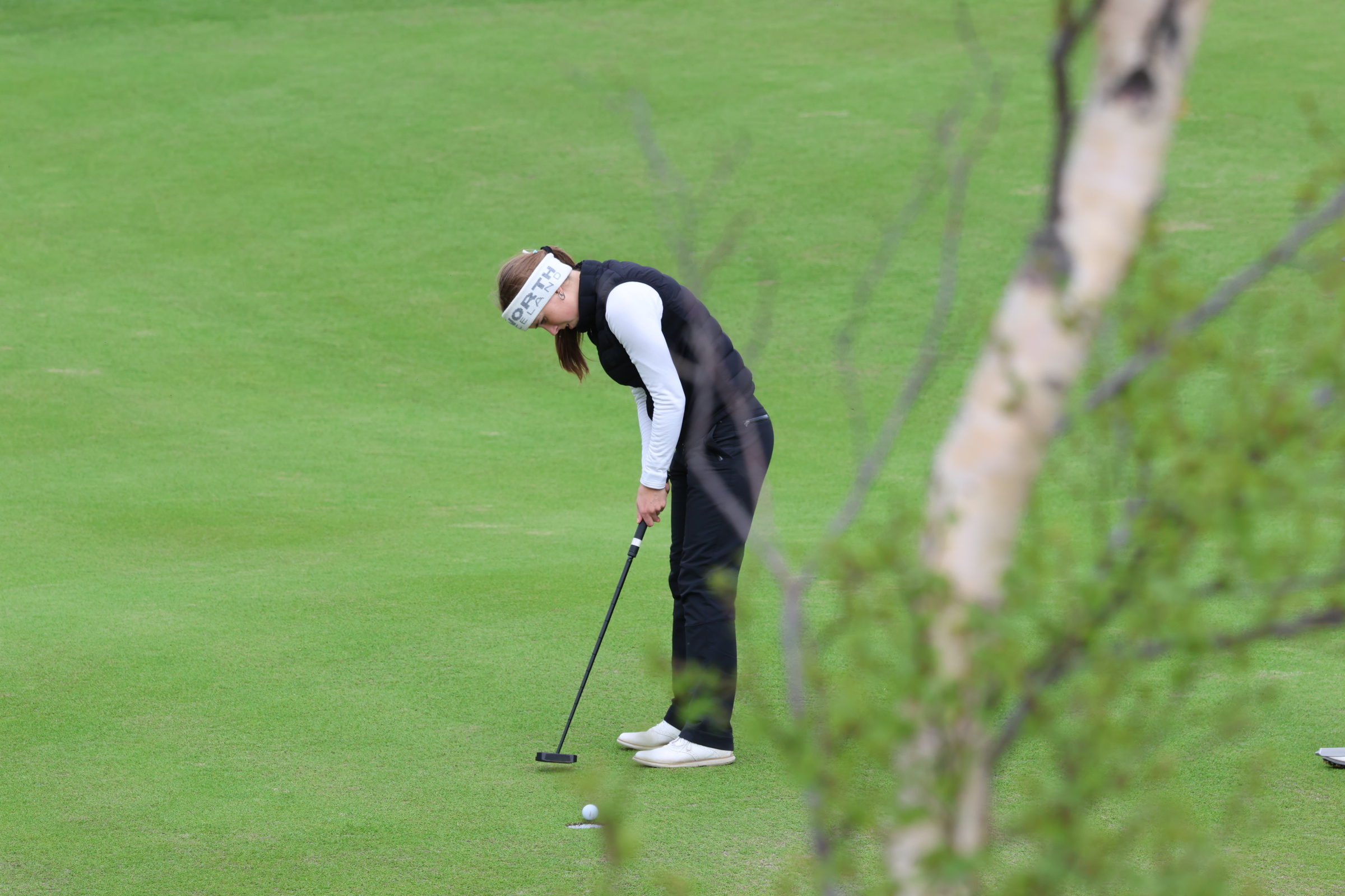 Golfer preparing a putt on a green, wearing a white long-sleeve top and black vest, headband on the forehead.