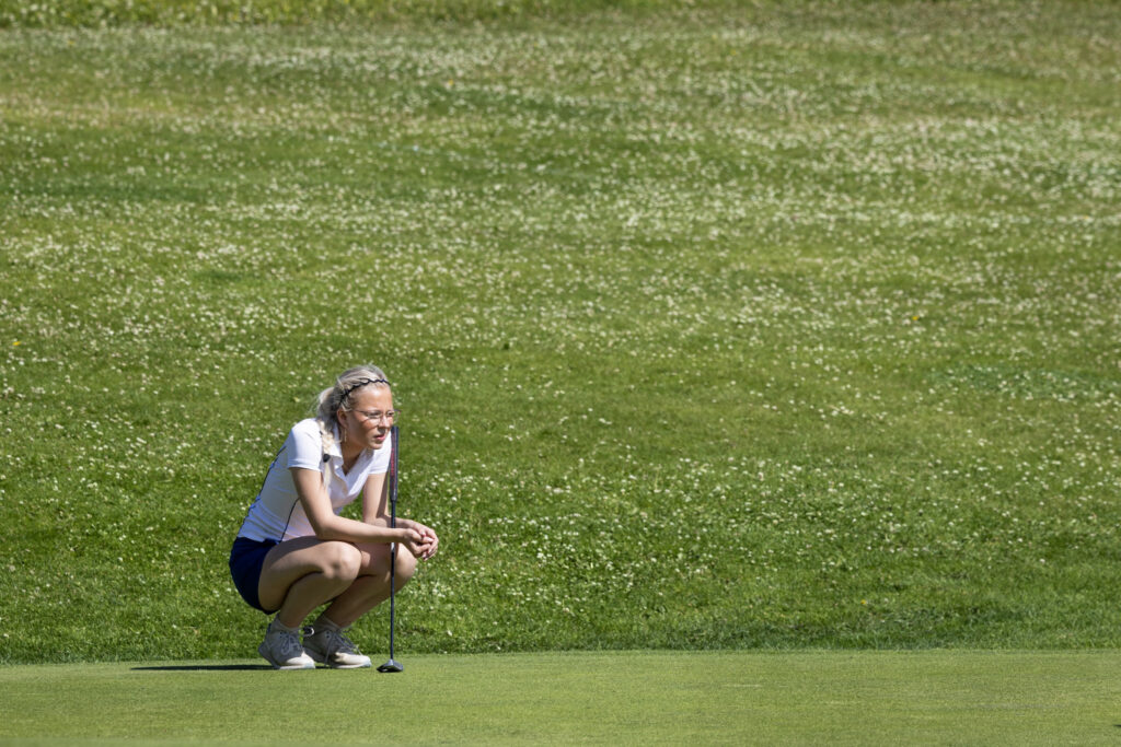 Woman crouching on a golf green with a putter, lining up a putt on a sunny course.