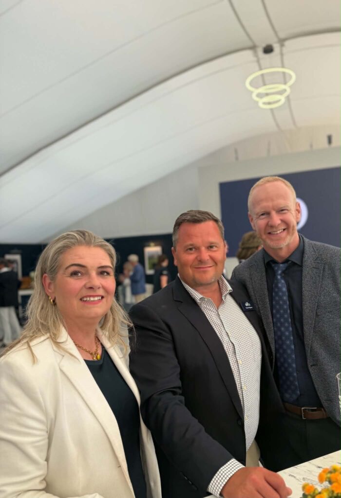 5df1c9fd-e8f9-4e78-9e3a-6f3cc56aa37f - Golfsamband Íslands Three adults smiling for a selfie at an indoor event; woman in a white blazer on the left, two men in suits nearby near a table with flowers.