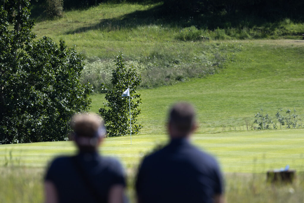 Blurred silhouettes of spectators on a sunny golf course with a flagstick on the green in the middle distance and lush fairway beyond.
