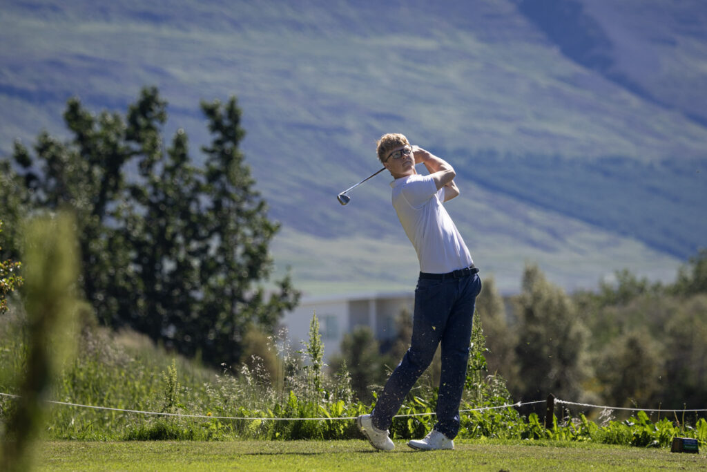 Golfer mid-swing on a sunny golf course with mountains in the background, wearing a white polo and dark pants.