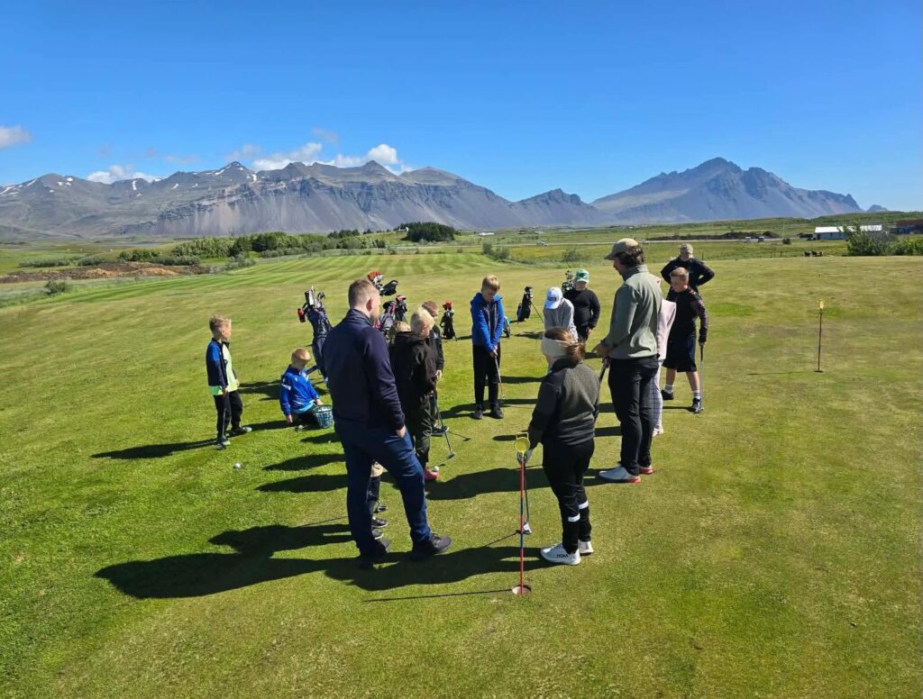730570525_18056531843769769_2948338444965147125_n 1 - Golfsamband Íslands Group of people, including children, gathered on a golf practice green as a instructor demonstrates a swing with golf clubs, mountains in the distance under a clear blue sky.