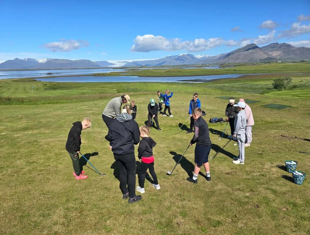 730713364_18056531825769769_5858988843816057624_n 1 - Golfsamband Íslands Group of people on a grassy hill practicing golf together, with a lake and snow-capped mountains in the background.