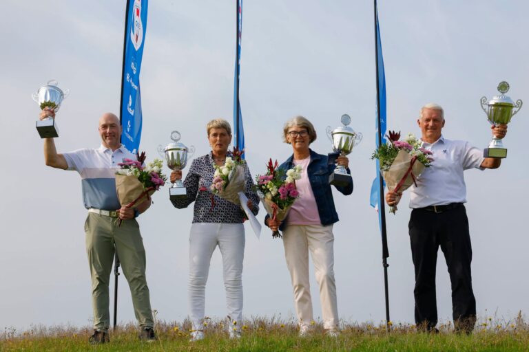 Five adults stand outdoors on a grassy field, each holding a trophy and a bouquet, posing for a victory photo with blue banners in the background.