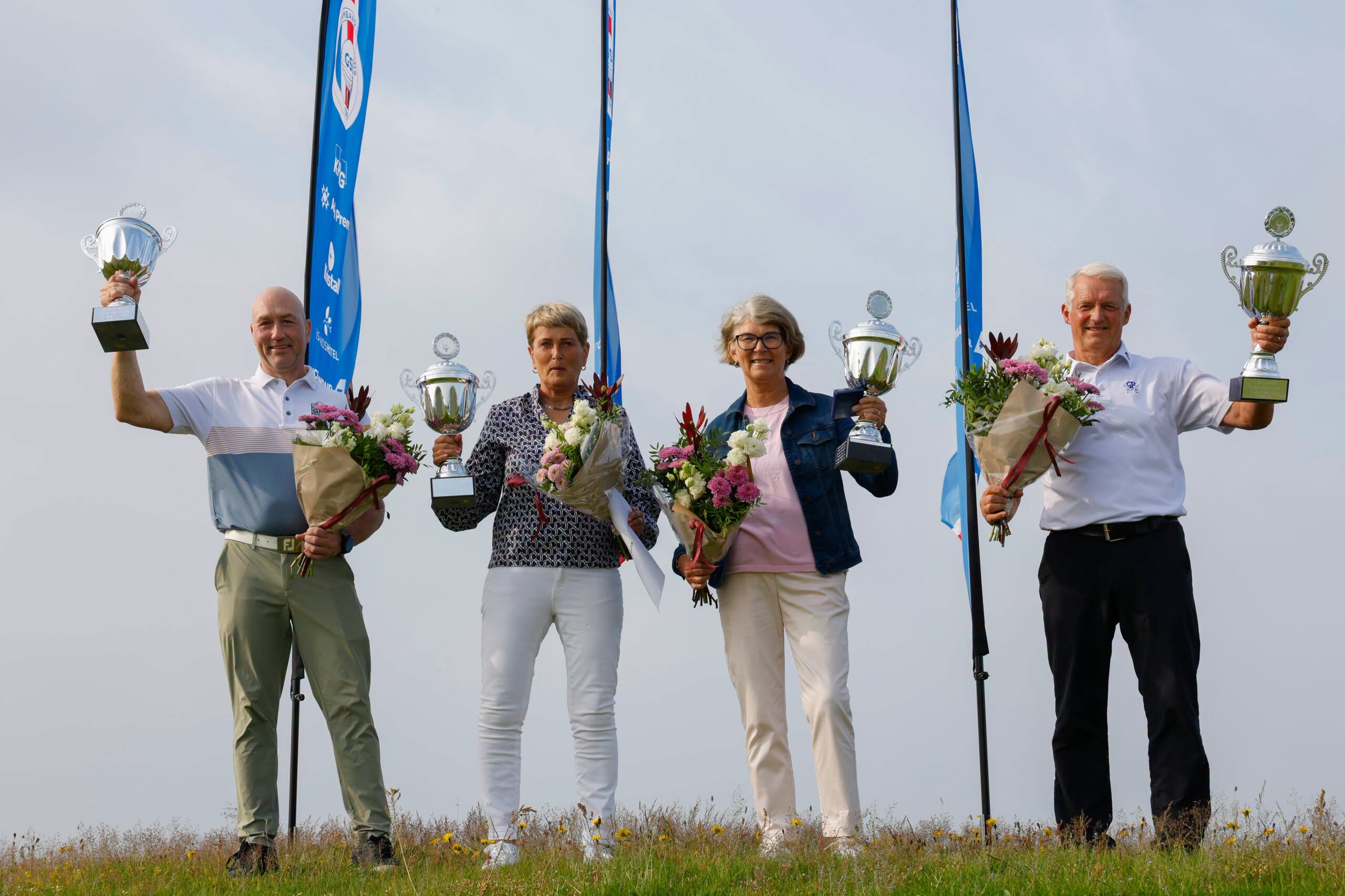 Five adults stand outdoors on a grassy field, each holding a trophy and a bouquet, posing for a victory photo with blue banners in the background.