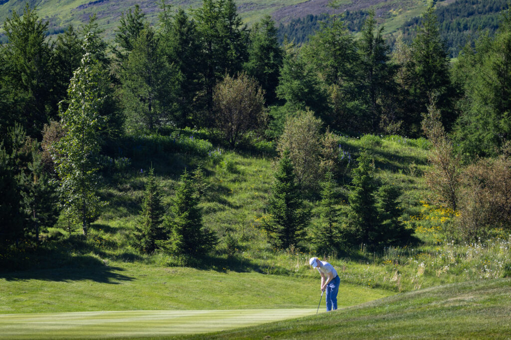 Golfer in blue pants bending to putt on a green with a forested hillside in the background.