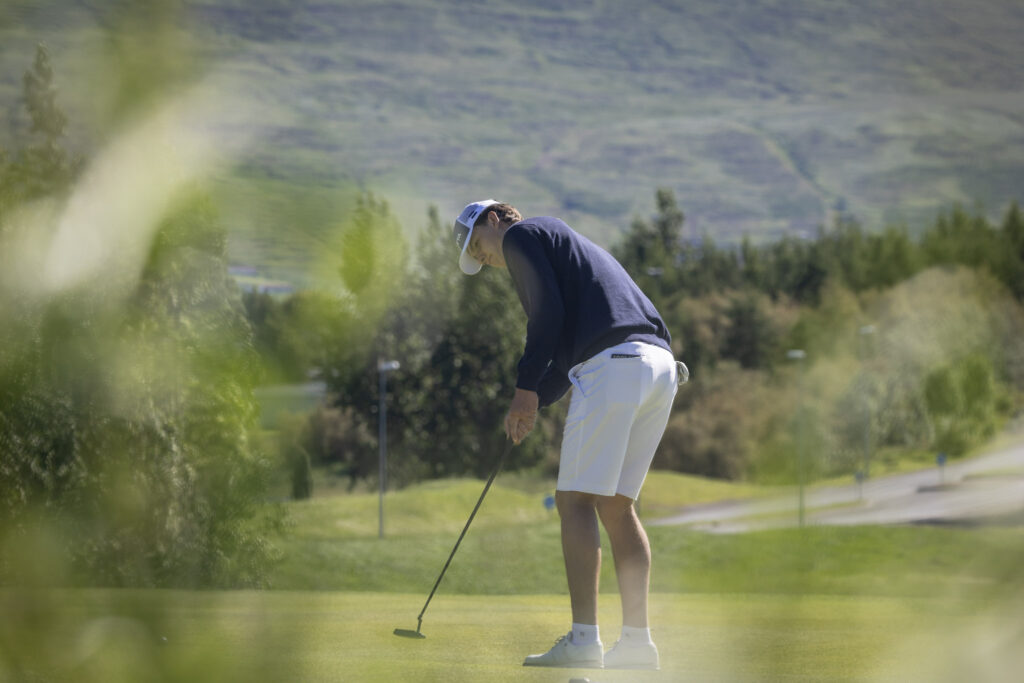 Golfer bending over to putt on a green with a hilly landscape in the background.