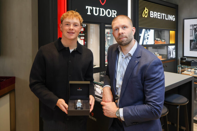 Two men stand at a watch counter; one presents a Tudor watch in an open box to the camera, while the other watches. Behind them are Tudor and Breitling displays in a store.","A salesperson in black and a man in a blue blazer pose with a Tudor watch box in a jewelry shop displaying Tudor and Breitling signs.","Man in a black shirt shows a Tudor wristwatch in its box to the camera, beside a colleague in a blue suit in a luxury watch shop.","Close-up view of a Tudor watch being presented in a black box, with store branding and other watches in the background.","Two men at a luxury watch shop; one holds an opened Tudor watch box showing a blue-faced timepiece, with brand displays in the background.