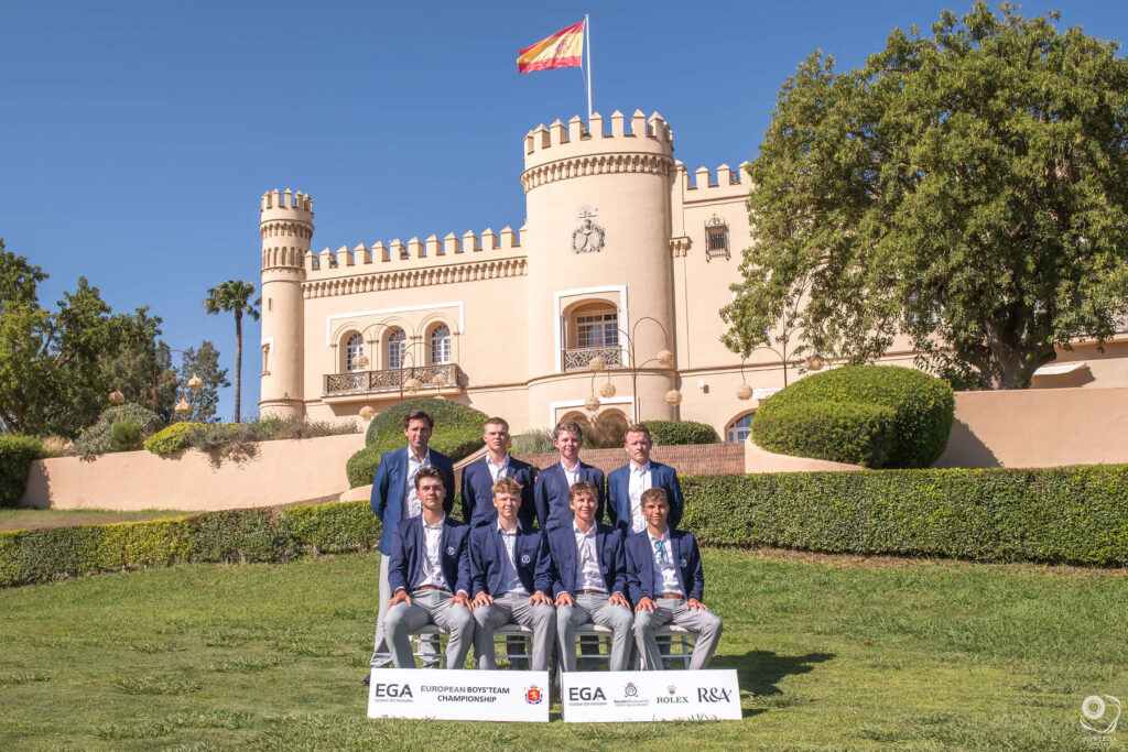 ETBCSortegafoto-213 - Golfsamband Íslands Group of young golfers in navy blazers posing on a lawn in front of a beige castle with a Spanish flag nearby.