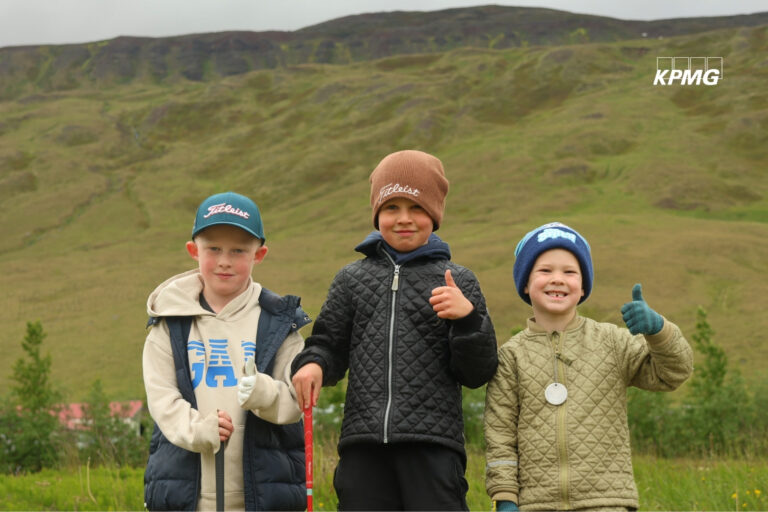 Three smiling boys in jackets and hats stand outdoors on a grassy hillside, thumbs up for a group photo with a red walking stick nearby. KPMG watermark in the corner.