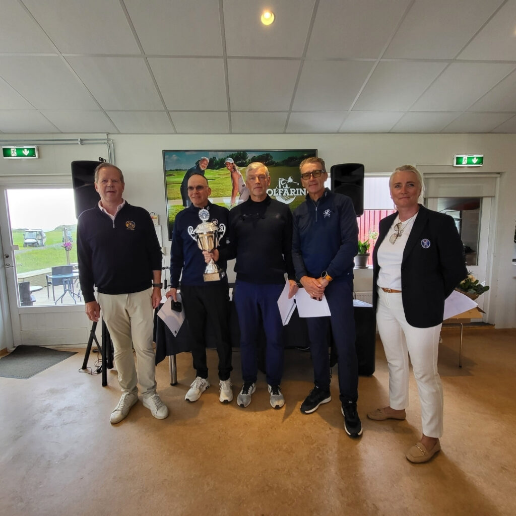 Group of five people in a golf clubroom posing with a large trophy and papers, celebrating an award ceremony.