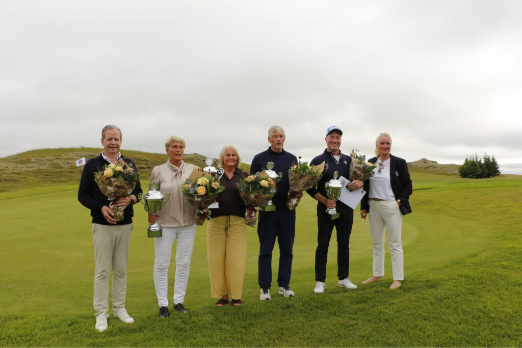 Group of seven people on a golf course holding bouquets and large trophies, posing for a winner’s photo.