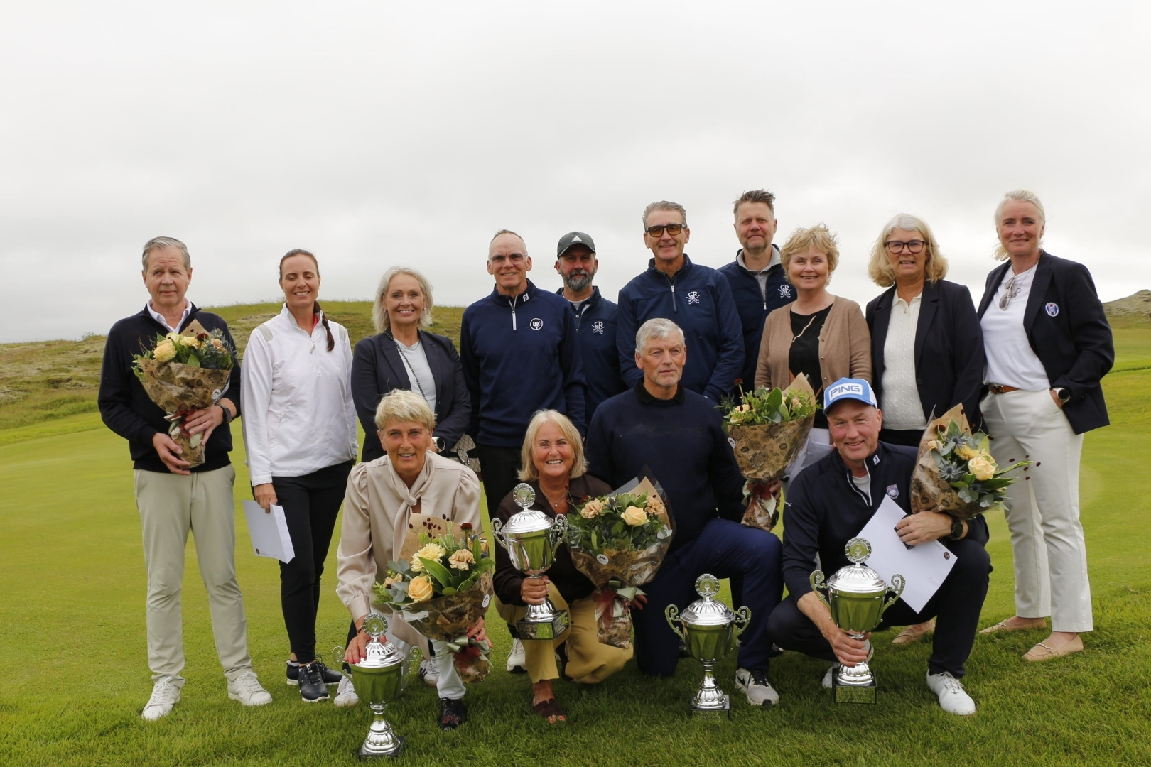 Group of twelve adults posing on a golf course with trophies and flower bouquets after a tournament.