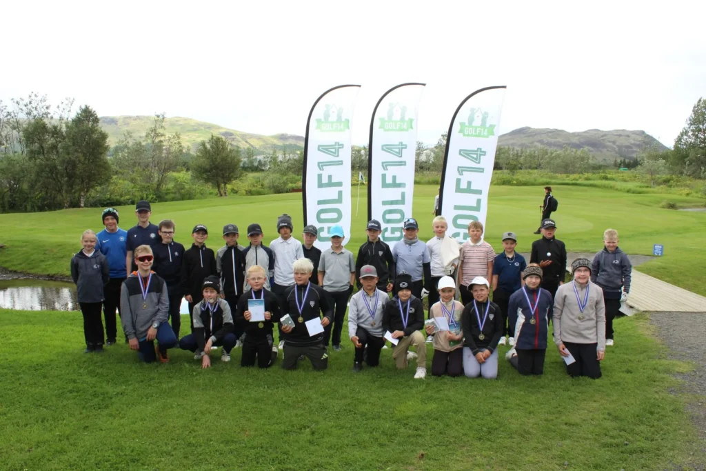 Group of children posing with medals on a green golf course, flags reading GOLF4 in the background.