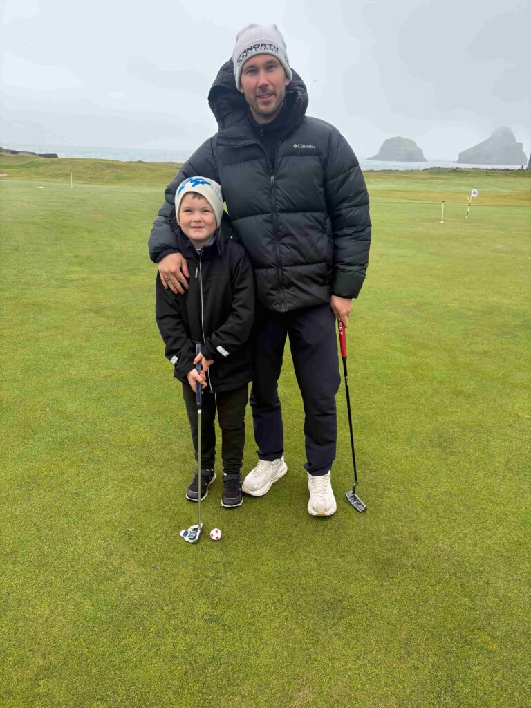 IMG_2786 - Golfsamband Íslands Man and a young boy stand together on a golf green, each holding a putter, with a hole and ball in the foreground and the sea and rocky outcrops in the background.