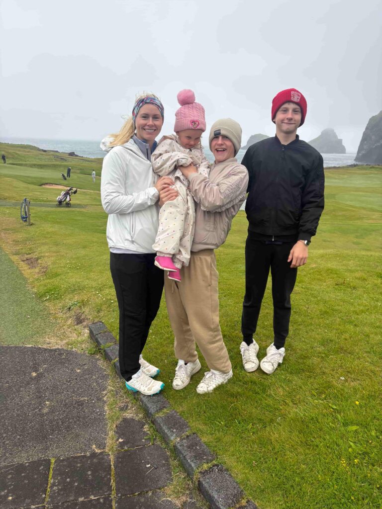 IMG_2787 - Golfsamband Íslands Smiling family of four posing on a coastal golf course, with rocky sea cliffs in the background on a cloudy day.