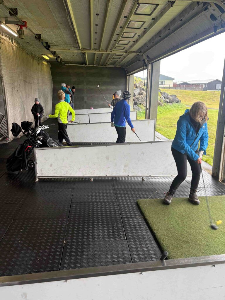 IMG_2788 - Golfsamband Íslands Group of people practicing golf indoors on matting; several players swing clubs near a dark back wall, with a coach or teammate observing in bright jackets.