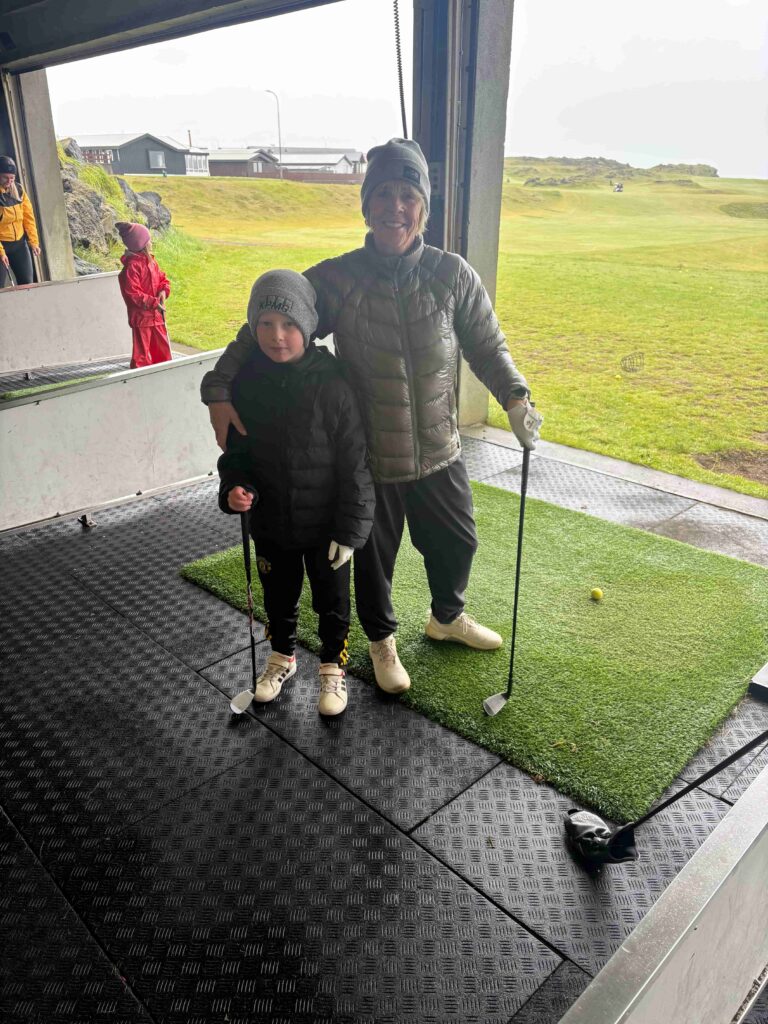 IMG_2789 - Golfsamband Íslands A woman and a young boy pose with golf clubs inside a covered driving range, outdoors on a grassy field.