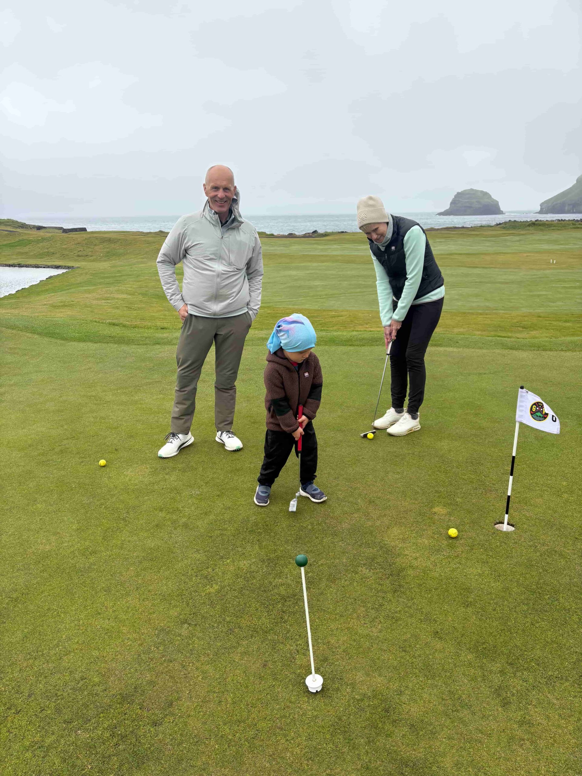 A man smiles beside a woman putting on a seaside golf green with a child watching nearby.