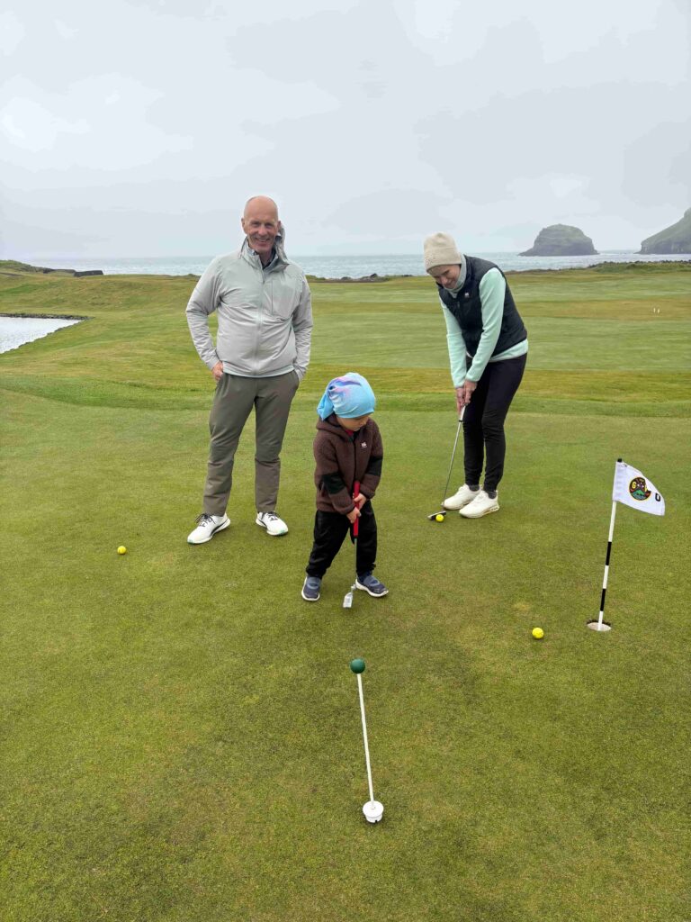 IMG_2790 - Golfsamband Íslands A man and a woman guide a young child putting on a green golf course by the coast, with the sea and rocky outcrops in the background.