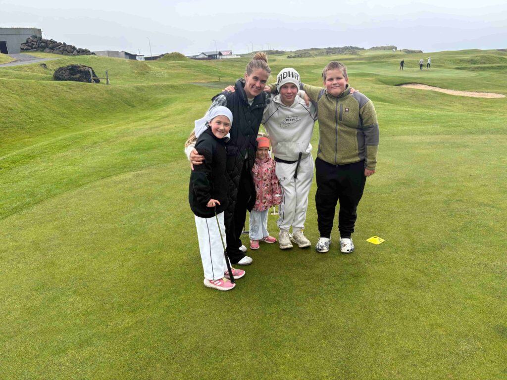 IMG_2791 - Golfsamband Íslands Five children stand together on a golf course green, smiling, with clubs in hand and a scenic course behind them.