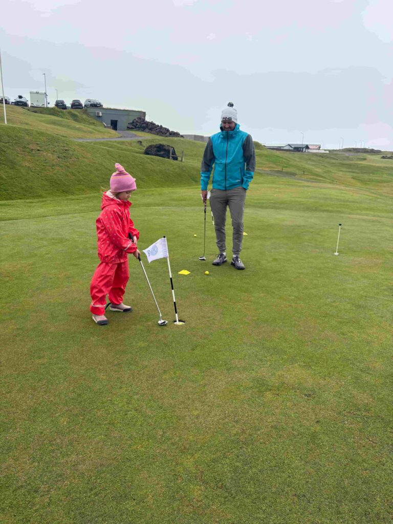 IMG_2792 - Golfsamband Íslands Man in a blue jacket and gray pants watches a child in a pink snowsuit practicing putts on a golf green.