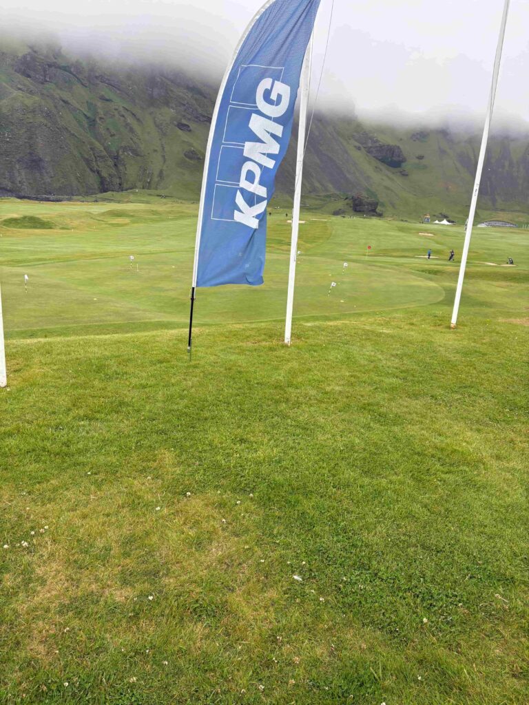 IMG_2793 - Golfsamband Íslands Blue flag banner with 'KPMG' on a green golf course, misty mountains in the background.