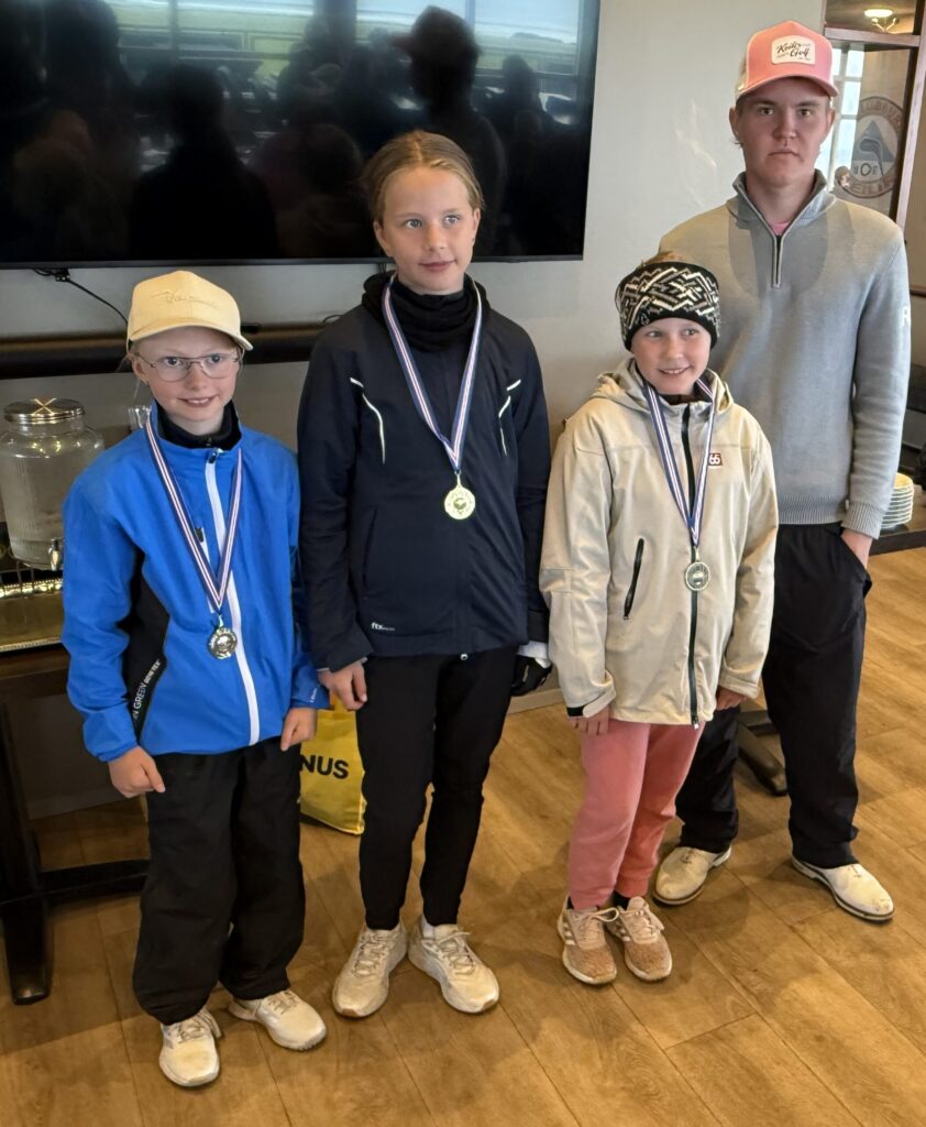 IMG_2953-scaled - Golfsamband Íslands Four young athletes wearing medals pose indoors for a group photo, with three standing in front and one taller person behind them, all in athletic jackets and sneakers.