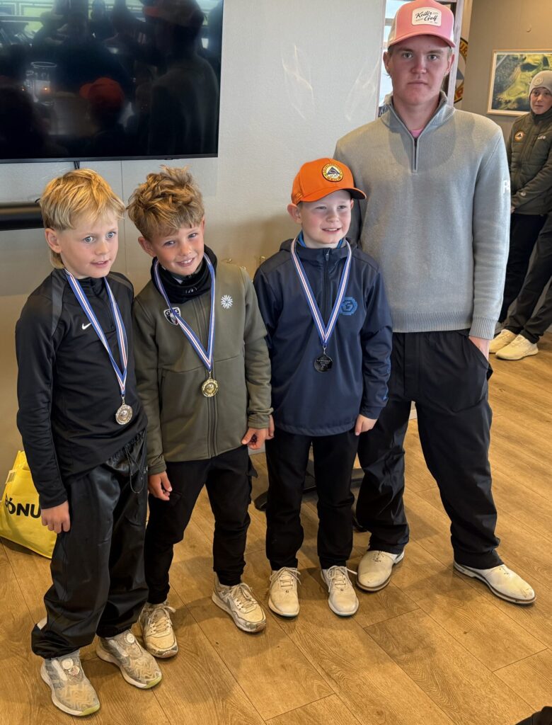IMG_2955-scaled - Golfsamband Íslands Four young boys with medals posing indoors beside a man in a gray sweater wearing caps and athletic outfits.
