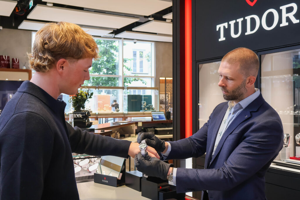 Customer in a dark jacket receives a wristwatch from a salesman at a Tudor showroom counter.