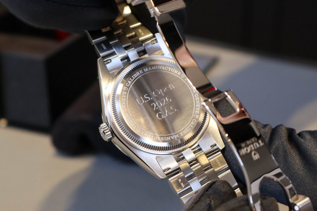 Close-up of a stainless steel wristwatch held by a gloved hand, showing the engraved back with 'U.S. Open 2026'.