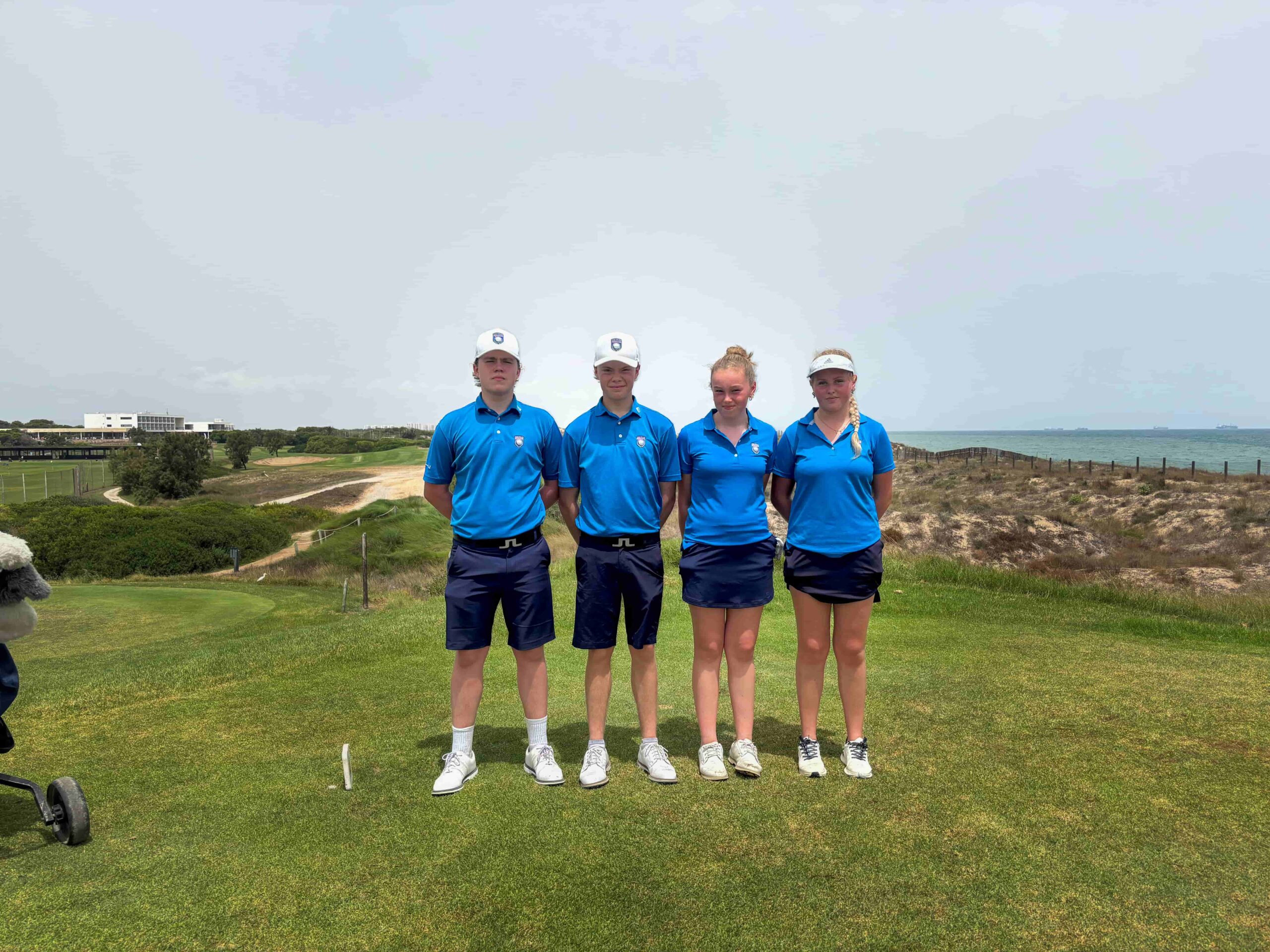 Four young golfers standing in a row on a sunny coastal golf course, wearing matching blue polo shirts and white caps.