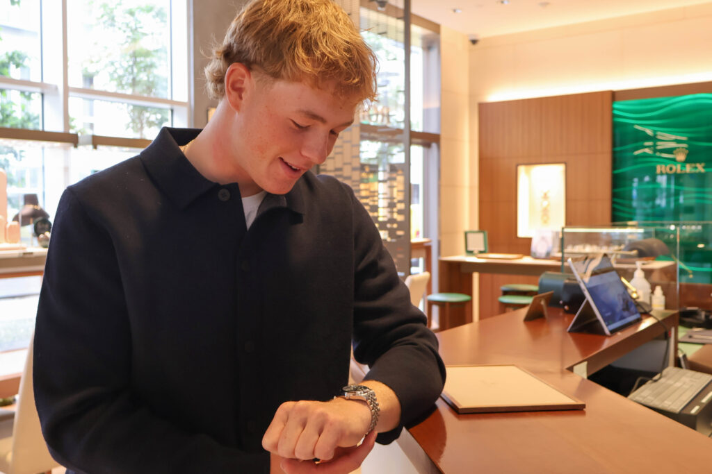 Man in a black polo leans over a wooden counter, looking down at his wristwatch in a showroom.