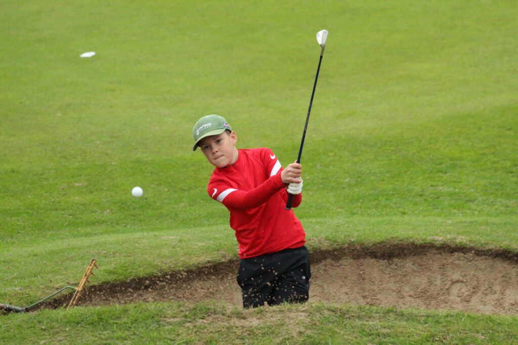 IMG_3579 - Golfsamband Íslands Young boy in a red shirt swinging a golf club on a grassy golf course.