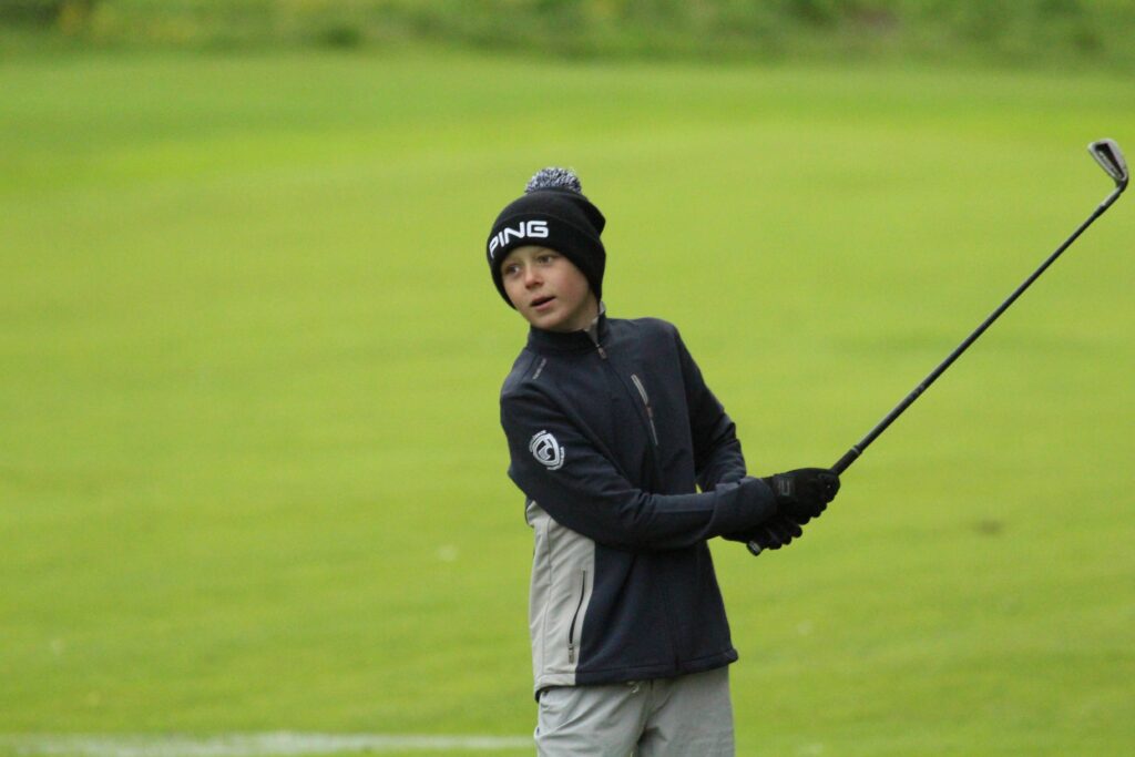 IMG_3588 - Golfsamband Íslands Young golfer in a black beanie and dark jacket swings a golf club on a green course.