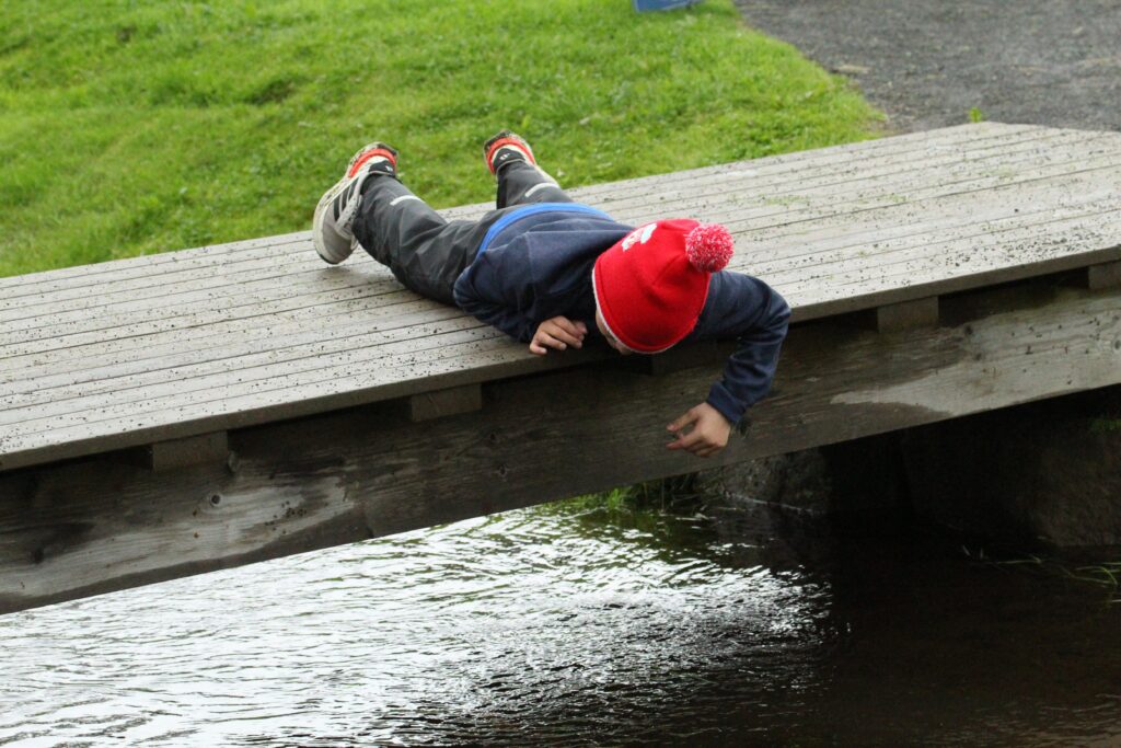 IMG_3598 - Golfsamband Íslands Child lying on a weathered wooden bridge, wearing a red knit hat with a pom-pom, feet up on the plank above the water.