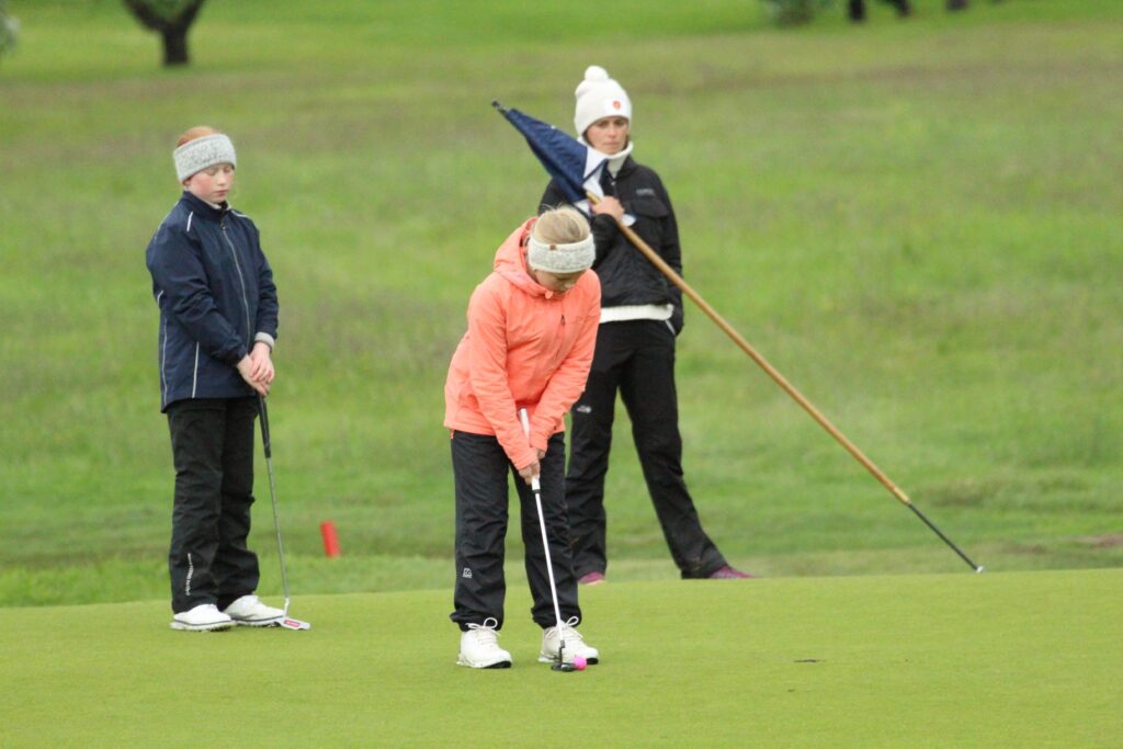 IMG_3603 - Golfsamband Íslands A woman in a coral jacket lines up a putt on the green while two other players stand nearby with their clubs.