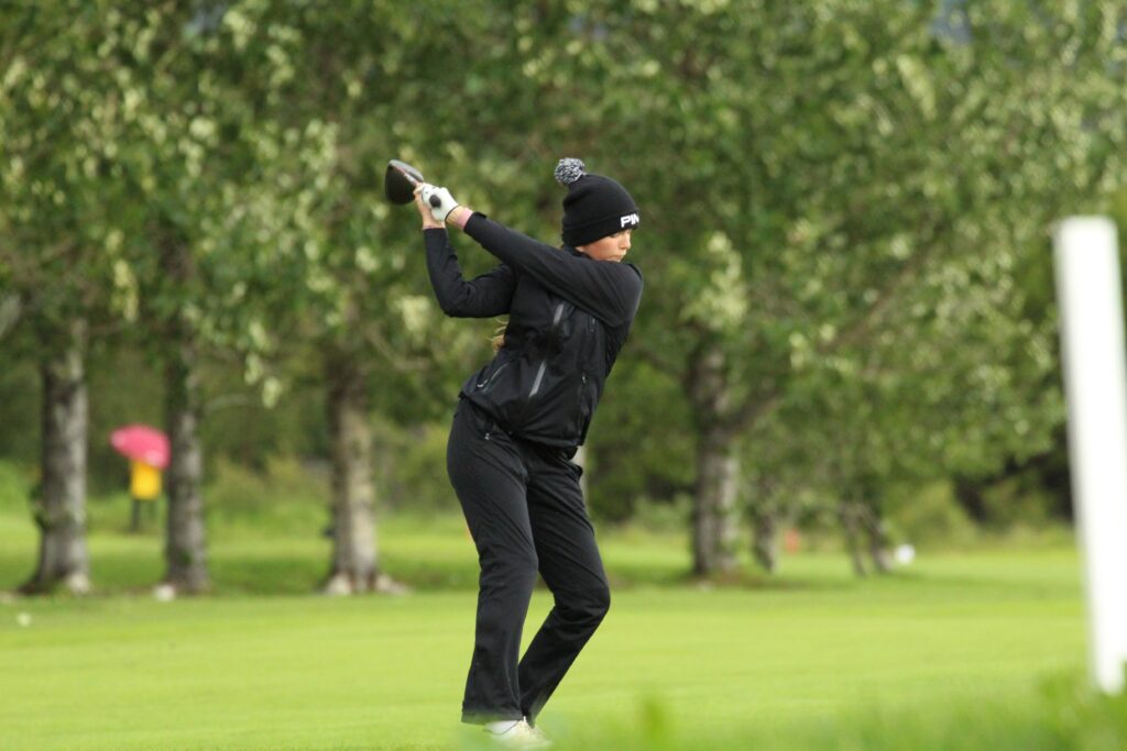 IMG_3626 - Golfsamband Íslands Person in black athletic wear swinging a golf club on a grassy field with trees in the background, mid-swing.