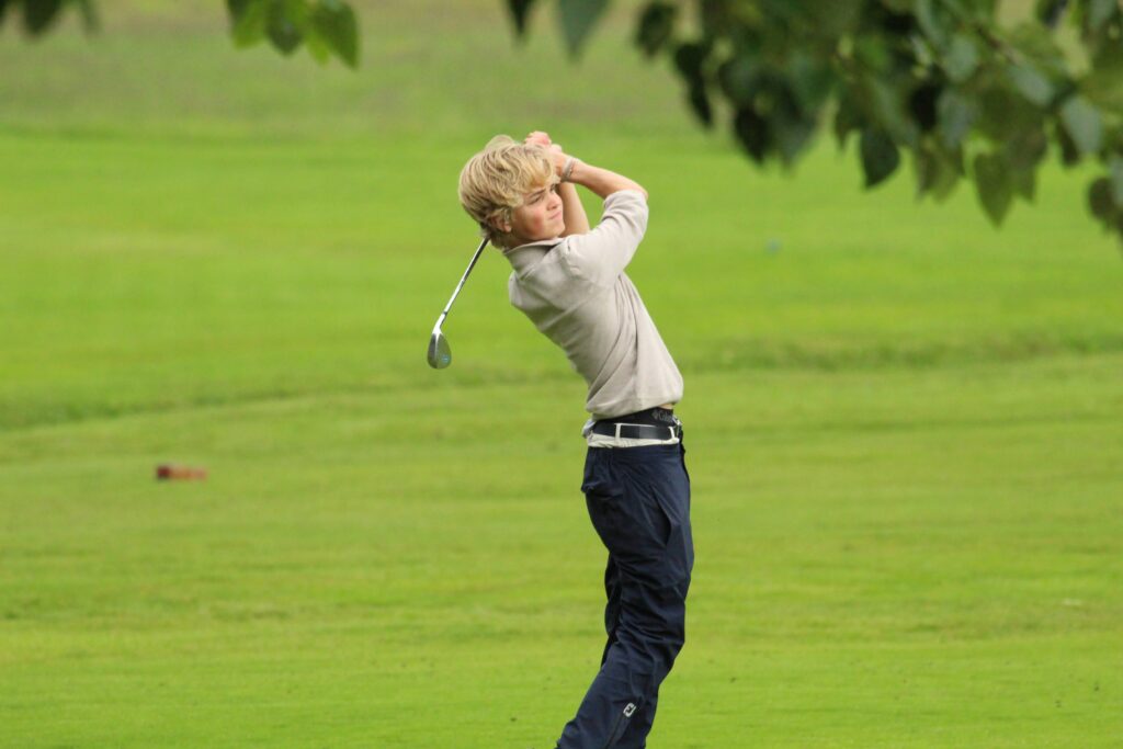 IMG_3648 - Golfsamband Íslands Young man in a light jacket mid-swing with a golf club on a grassy golf course