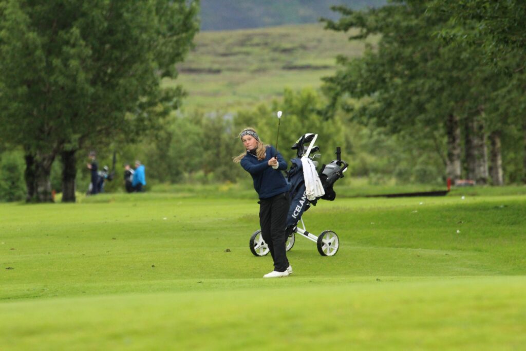 IMG_3653 - Golfsamband Íslands Female golfer mid-swing on a lush golf course, with a wheeled golf bag carrier nearby.
