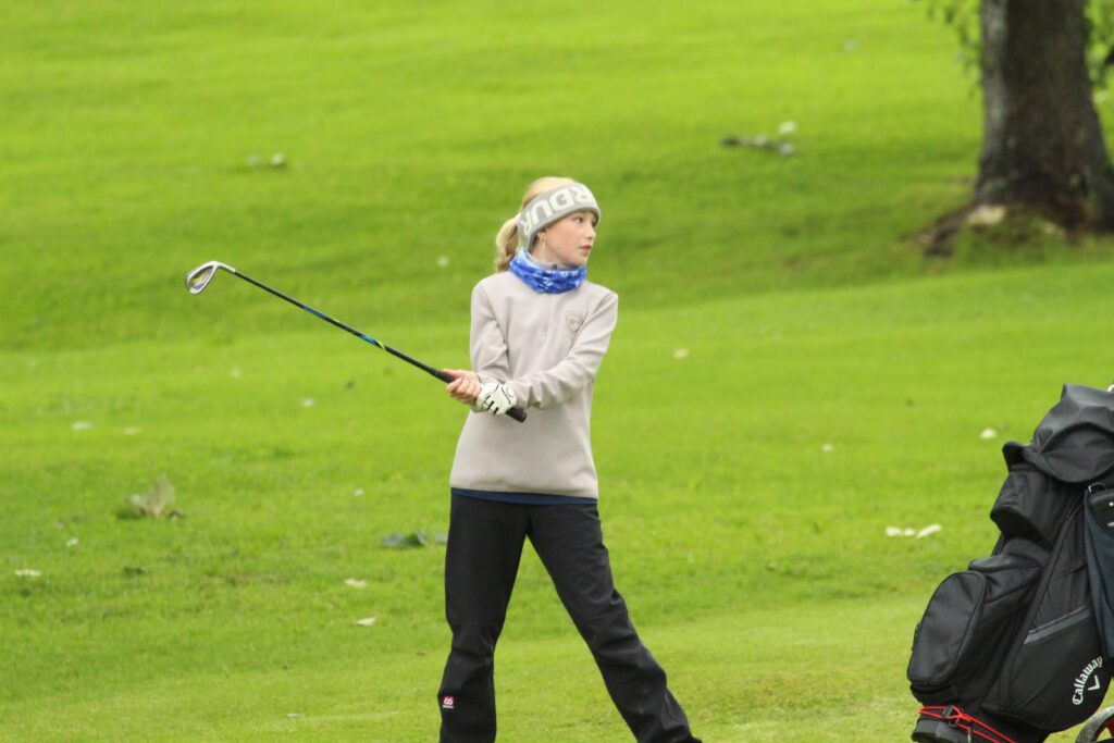 IMG_3670 - Golfsamband Íslands Girl in a beige jacket swinging a golf club on a grassy field, with a large golf bag nearby on the right side.